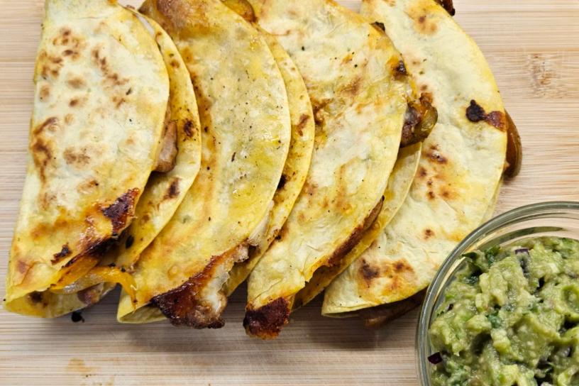 Crispy chicken tacos with vegetables on a wooden surface, with a small glass bowl of guacamole beside them.