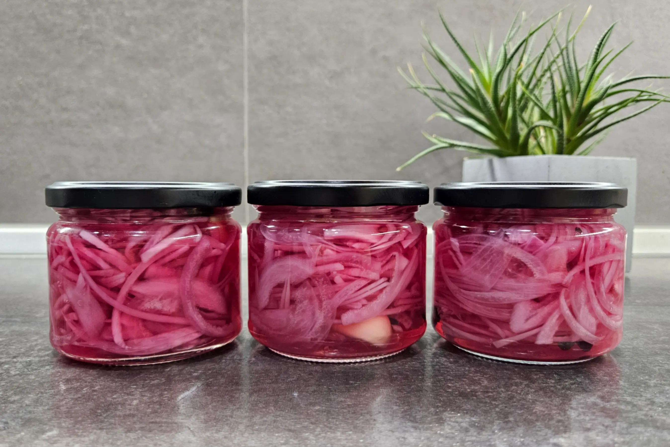 Три скляні банки з тонко нарізаною маринованою червоною цибулею в розсолі на темній поверхні, поруч сукулент у бетонному горщику.