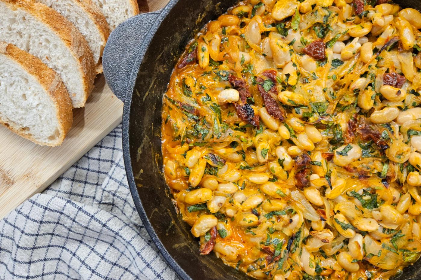 Квасоля канеліні у вершковому соусі з в’яленими томатами та зеленню в чавунній пательні, поруч нарізаний багет.