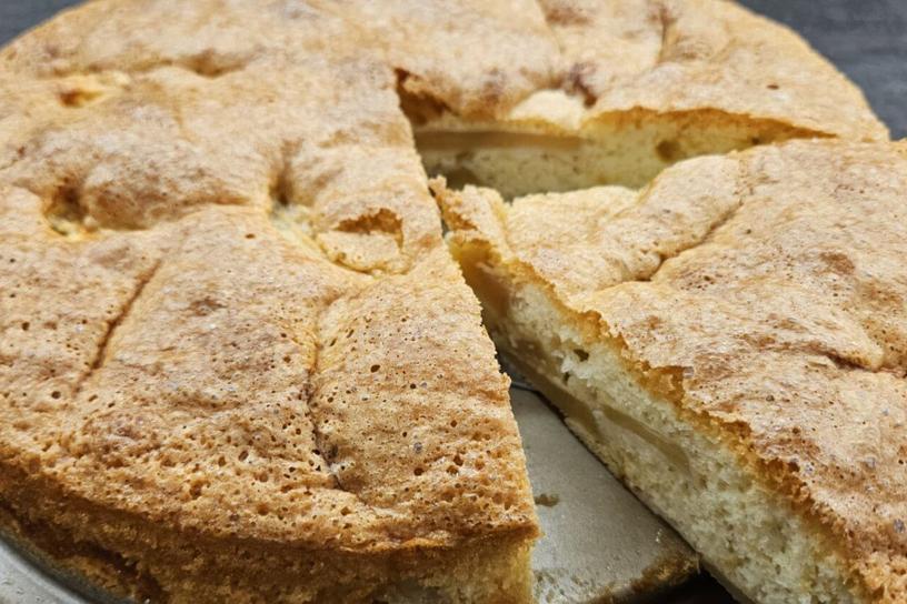 A golden-brown cake with a slice cut out, revealing its tender structure and a layer of apples.