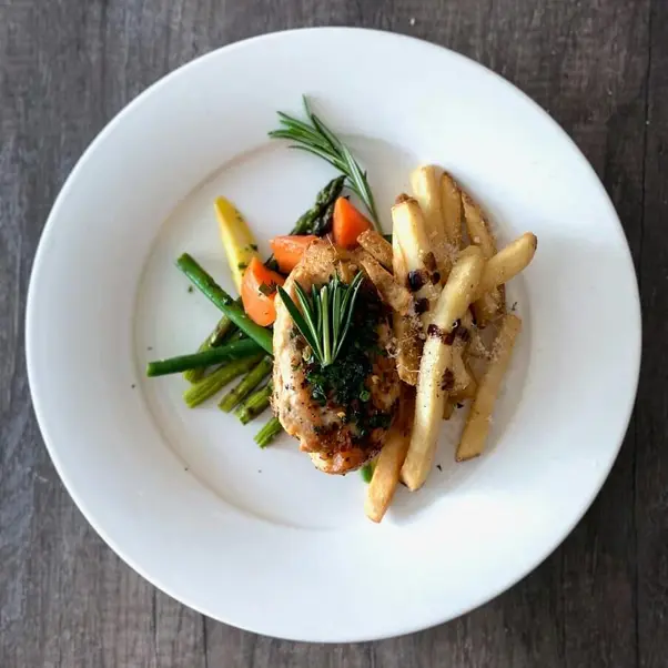 A white plate on a wooden table featuring grilled chicken, French fries, and vibrant vegetables.