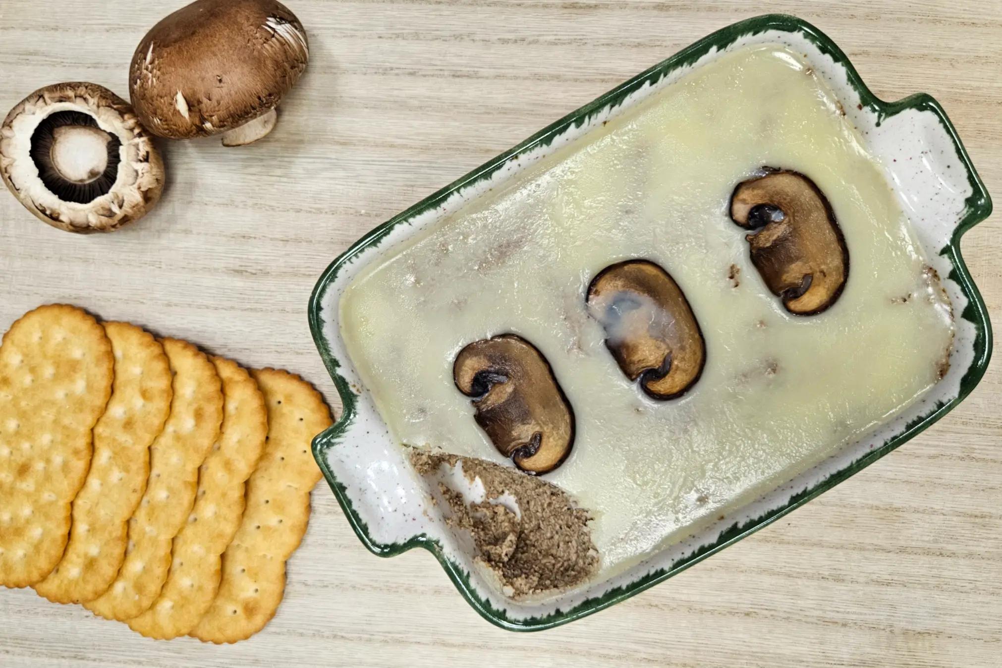 Кремовий грибний паштет у прямокутній формі, покритий вершковим маслом і трьома скибочками обсмажених грибів. Ліворуч видно кілька круглих солоних крекерів, а поруч — два свіжих гриба коричневого кольору.