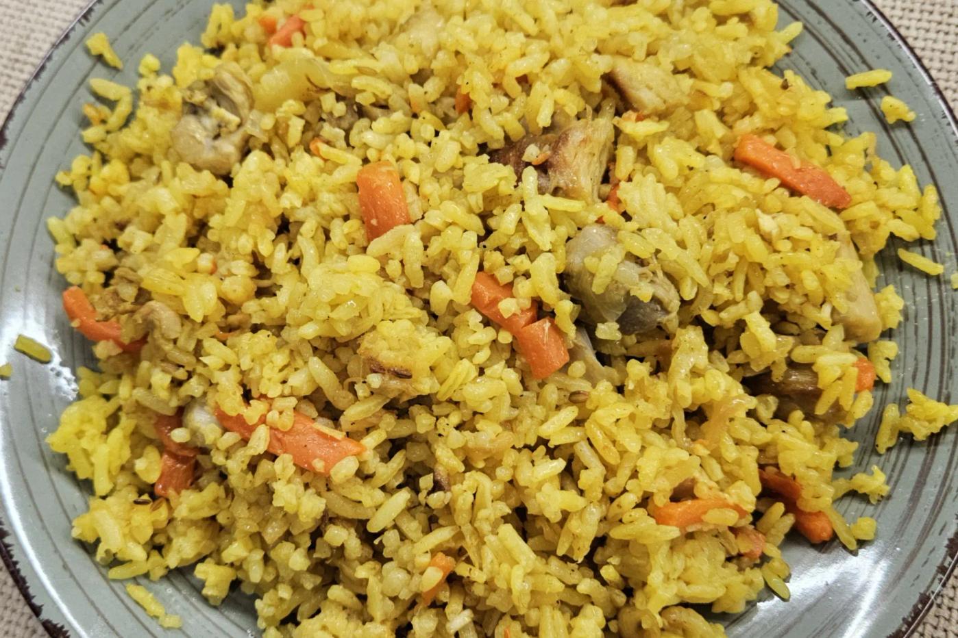 Fluffy chicken pilaf with carrot strips and spices on a gray‑green plate with a dark rim, set on a beige linen surface.