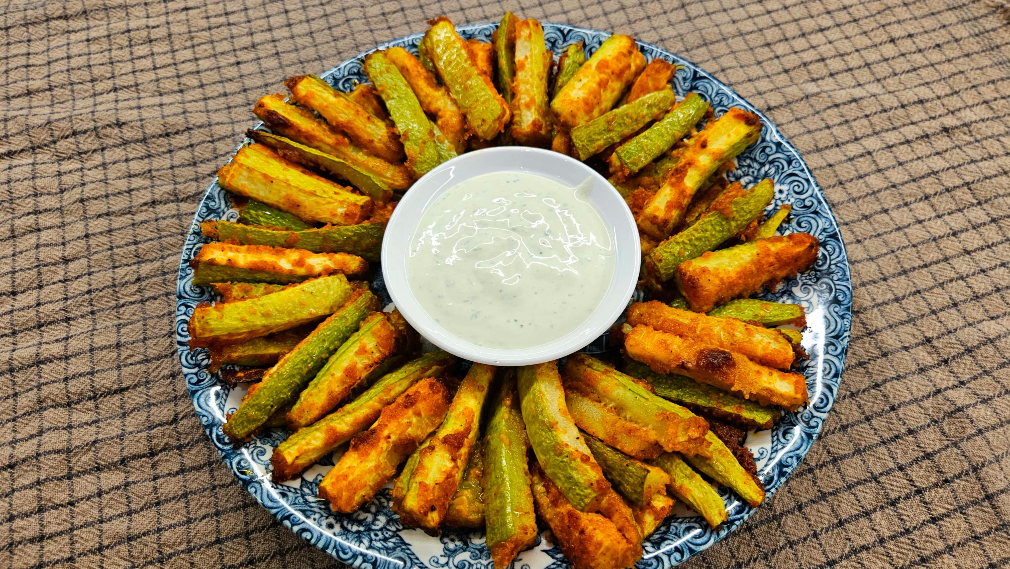 Crispy baked zucchini fries arranged in a circle on a serving plate around a small white bowl of garlic yogurt sauce.