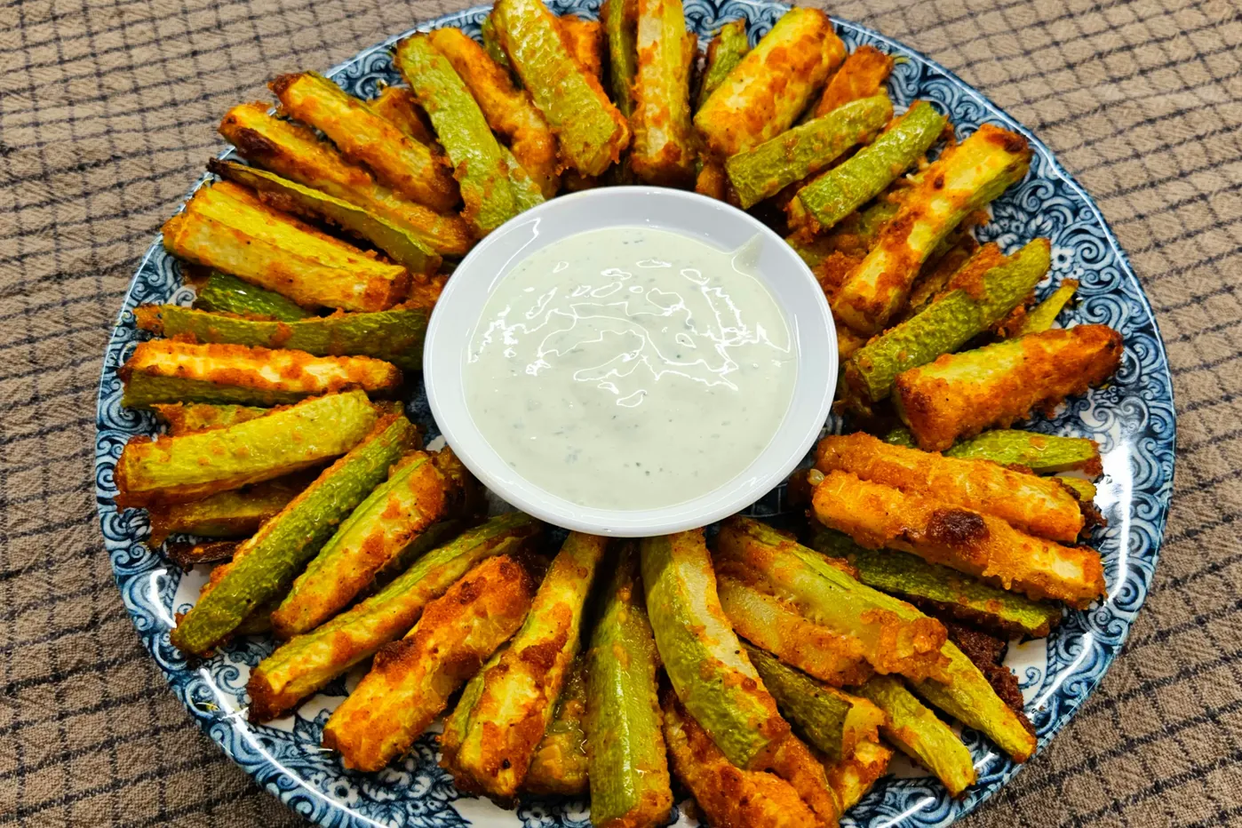 Crispy baked zucchini fries arranged in a circle on a serving plate around a small white bowl of garlic yogurt sauce.
