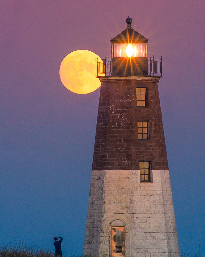 Large gold moon rising behind Point Judith lighthouse in Rhode Island with man standing taking a picture of it