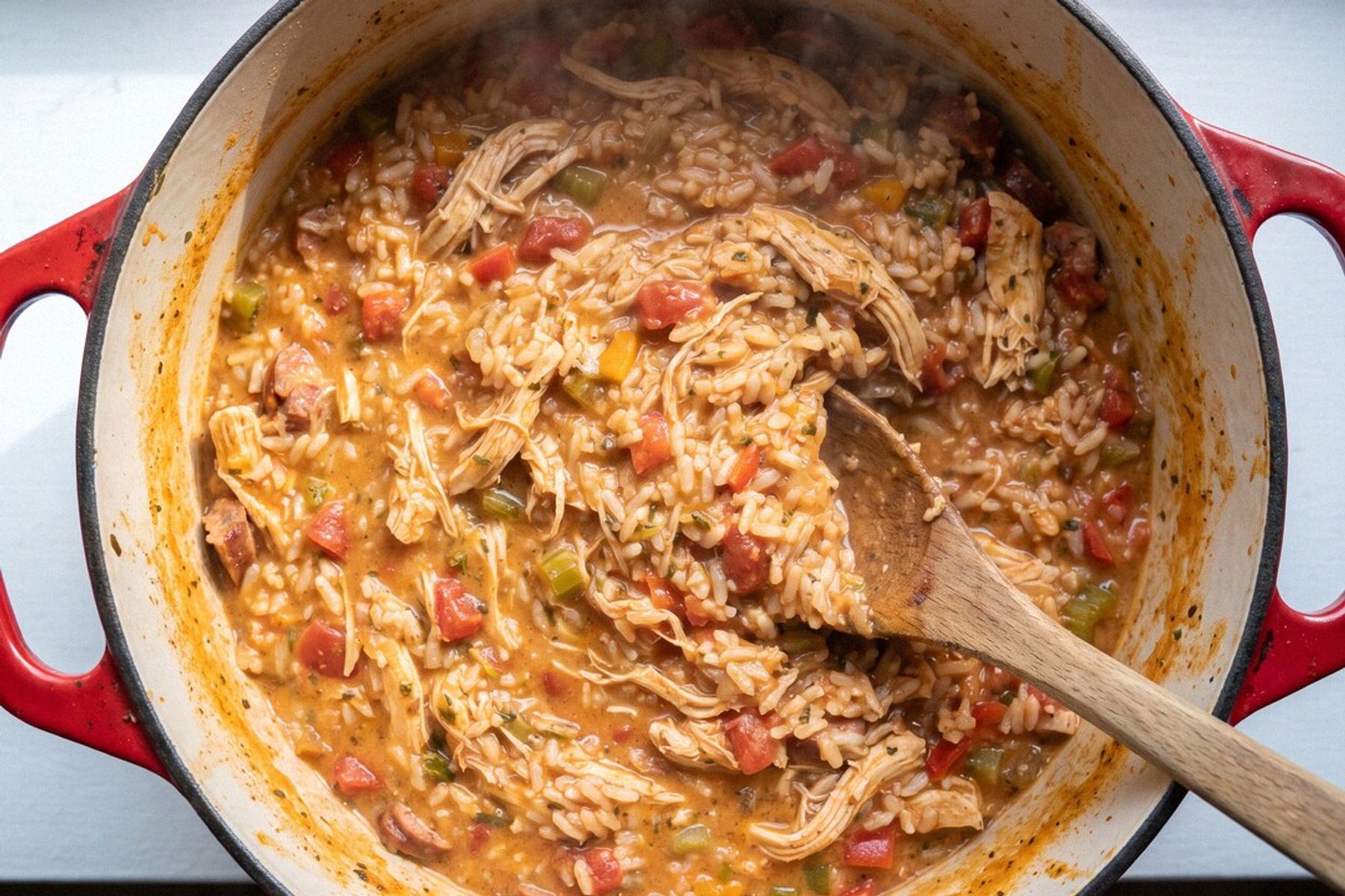 A red Dutch oven of big-batch jambalaya shot from overhead — creamy orange rice with shredded chicken, sausage and diced peppers, wooden spoon buried in the pot