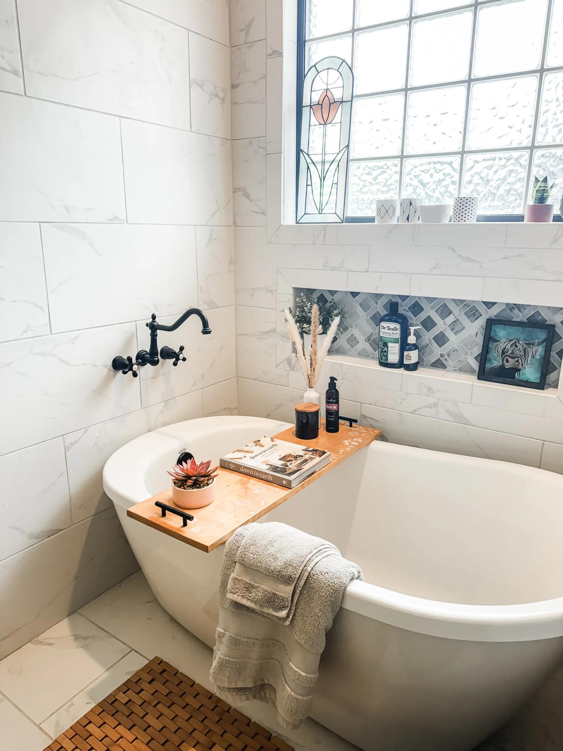 A finished wood bathtub tray styled across a soaking tub with a candle, plant, and book