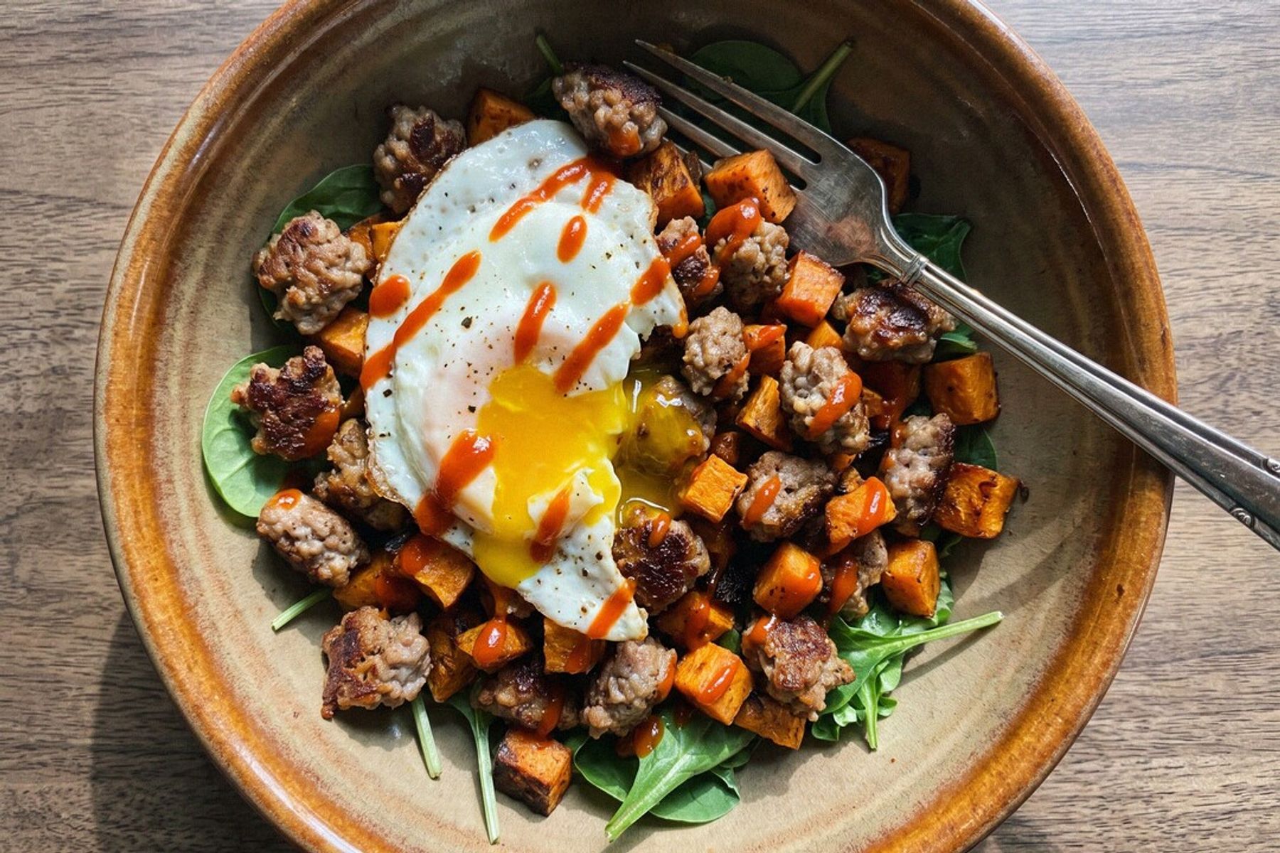 A rustic bowl of sausage and sweet potato hash over greens, topped with a fried egg with a runny yolk and a sriracha drizzle, shot from overhead
