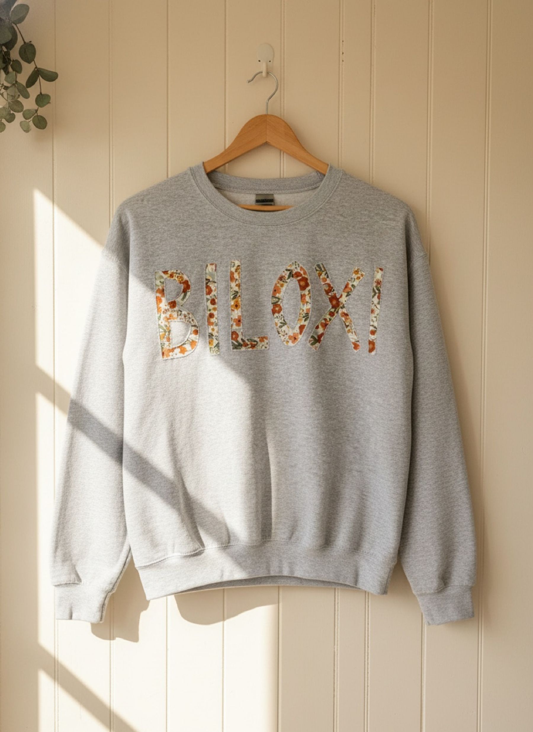 A grey crewneck sweatshirt with "BILOXI" spelled out in floral reverse appliqué, on a wooden hanger
