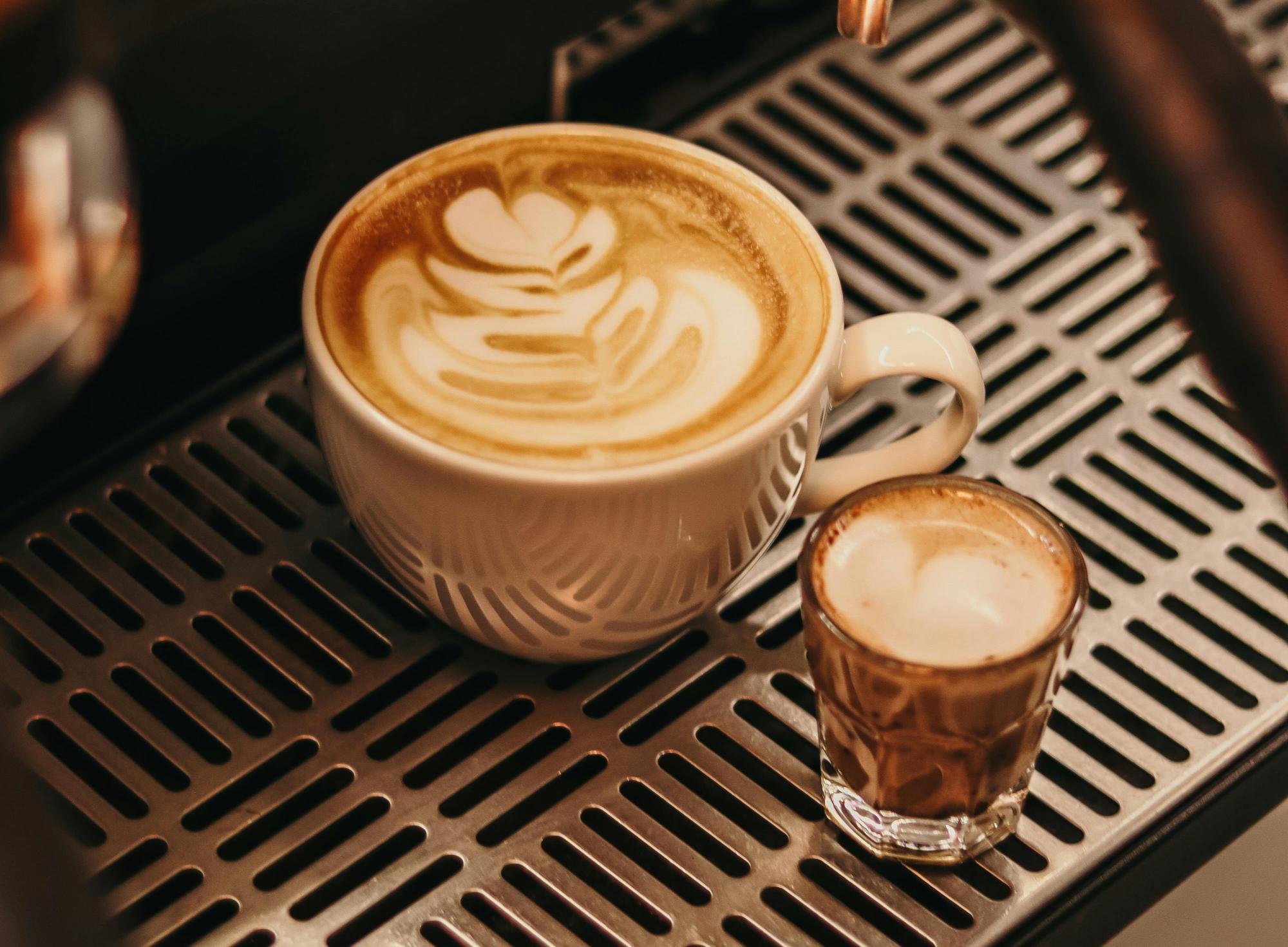 A latte with heart latte art and a small espresso shot on a metal grate.