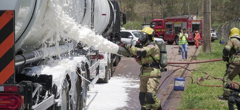 Bombeiro aplica espuma em caminhão-tanque durante simulado de emergência na BR-040, com viaturas e equipes ao fundo