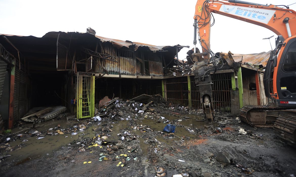 Escavadeira atua na demolição do pavilhão MFE-A incendiado na Ceagesp, em São Paulo, com escombros e estrutura metálica ao redor