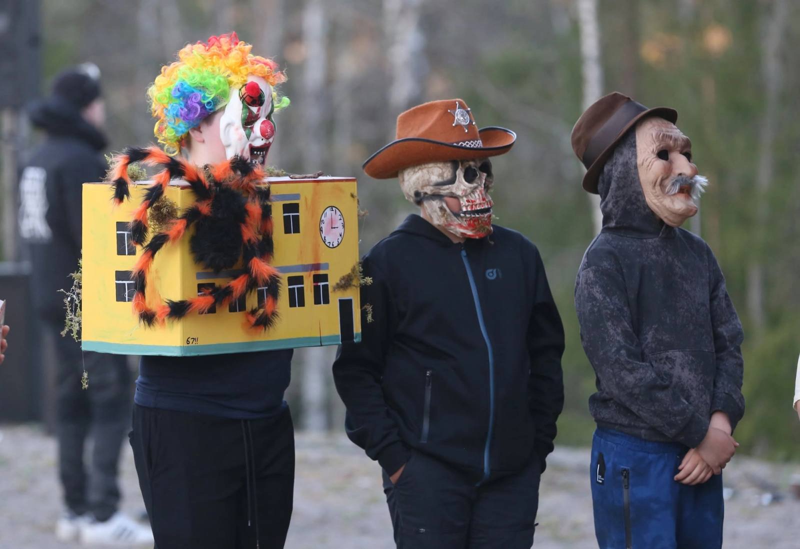 Tre valborrar i masker och utklädnad — en av dem bär en modell av ett gult hus.