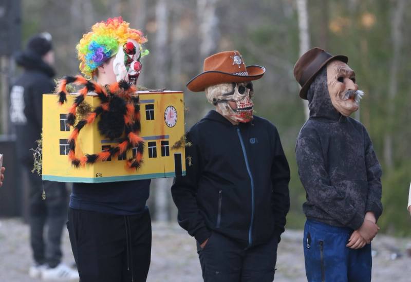 Tre valborrar i masker och utklädnad — en av dem bär en modell av ett gult hus.