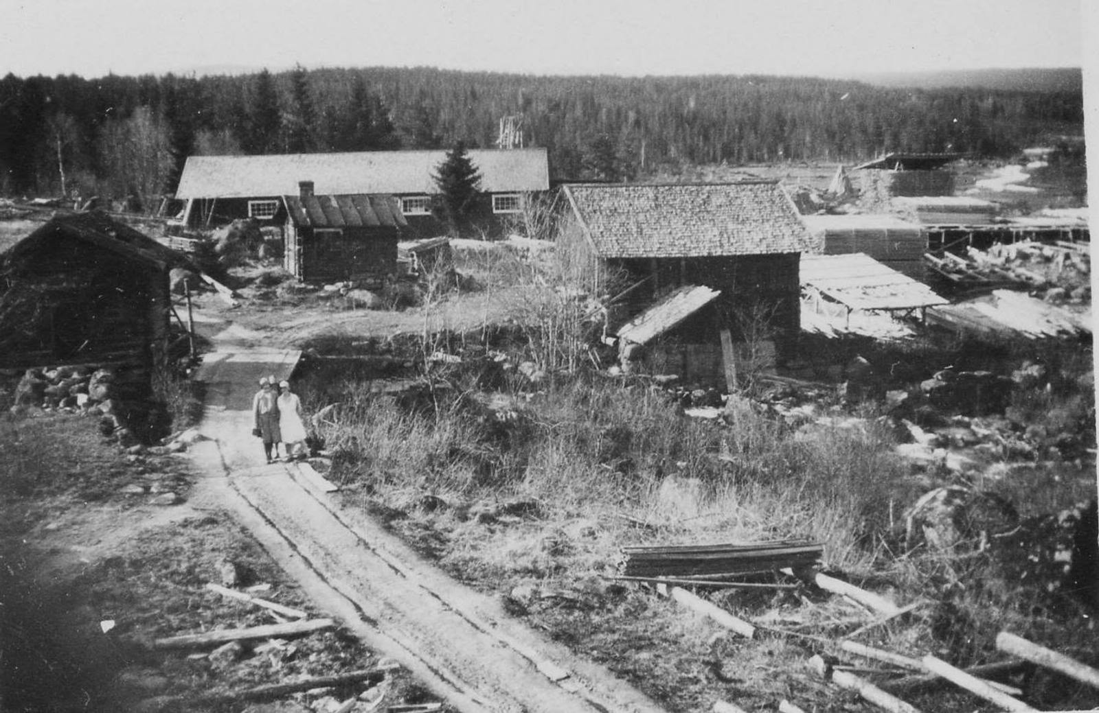 Gammalt svartvitt fotografi av byn: timrade hus, en grusväg och två barn på vägen.