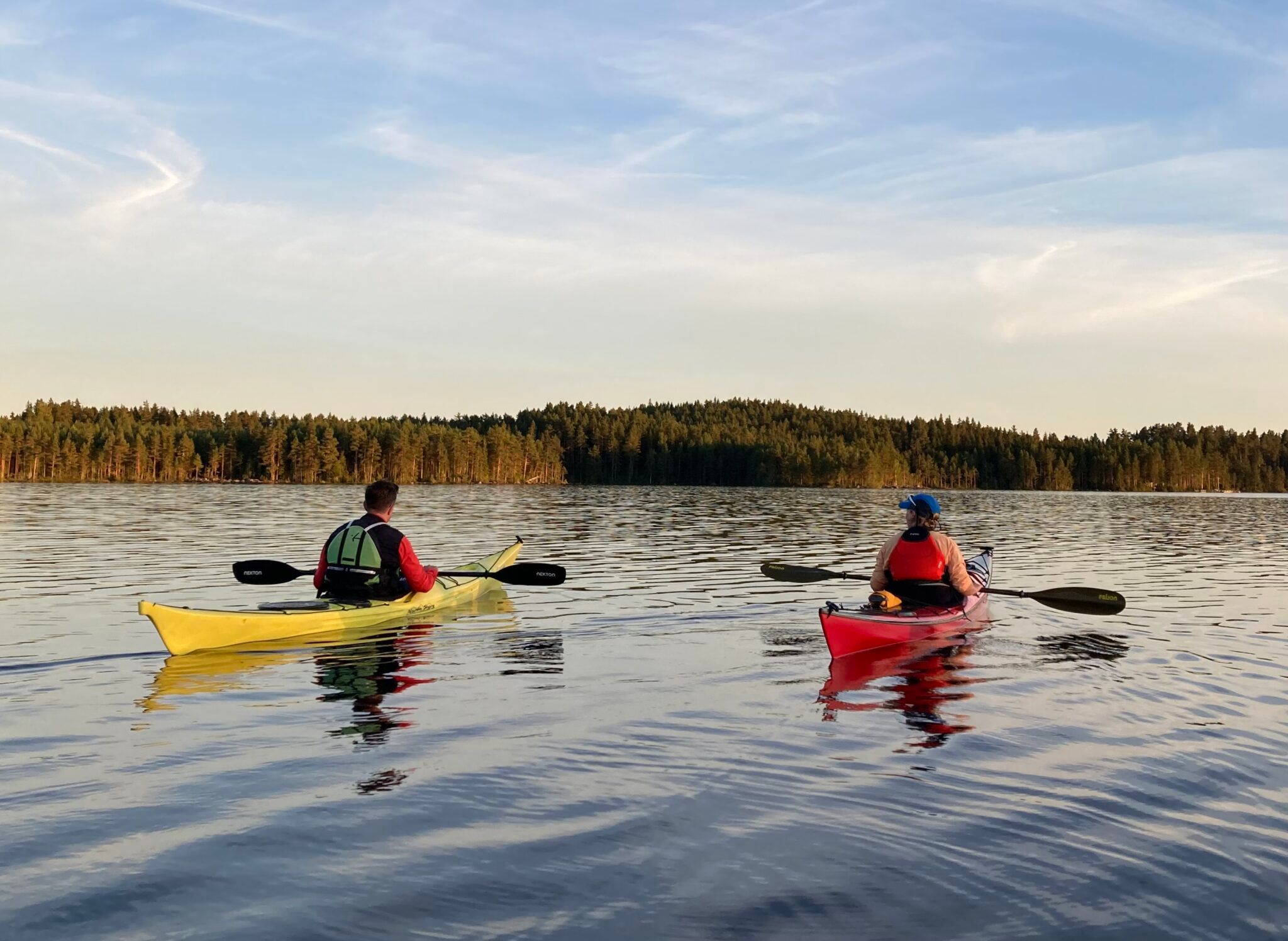 Två paddlare i gul och röd kajak på en blank sjö i kvällssol, med barrskog längs stranden.