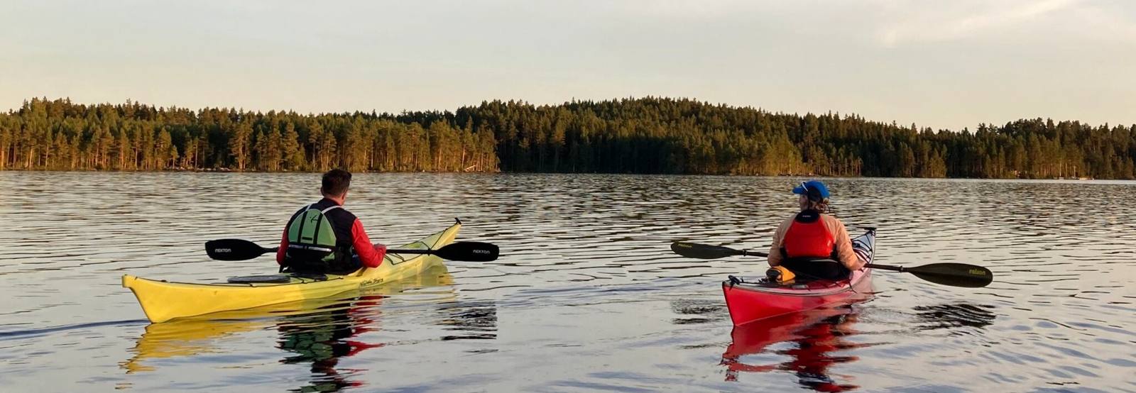 Två paddlare i gul och röd kajak på en blank sjö i kvällssol, med barrskog längs stranden.