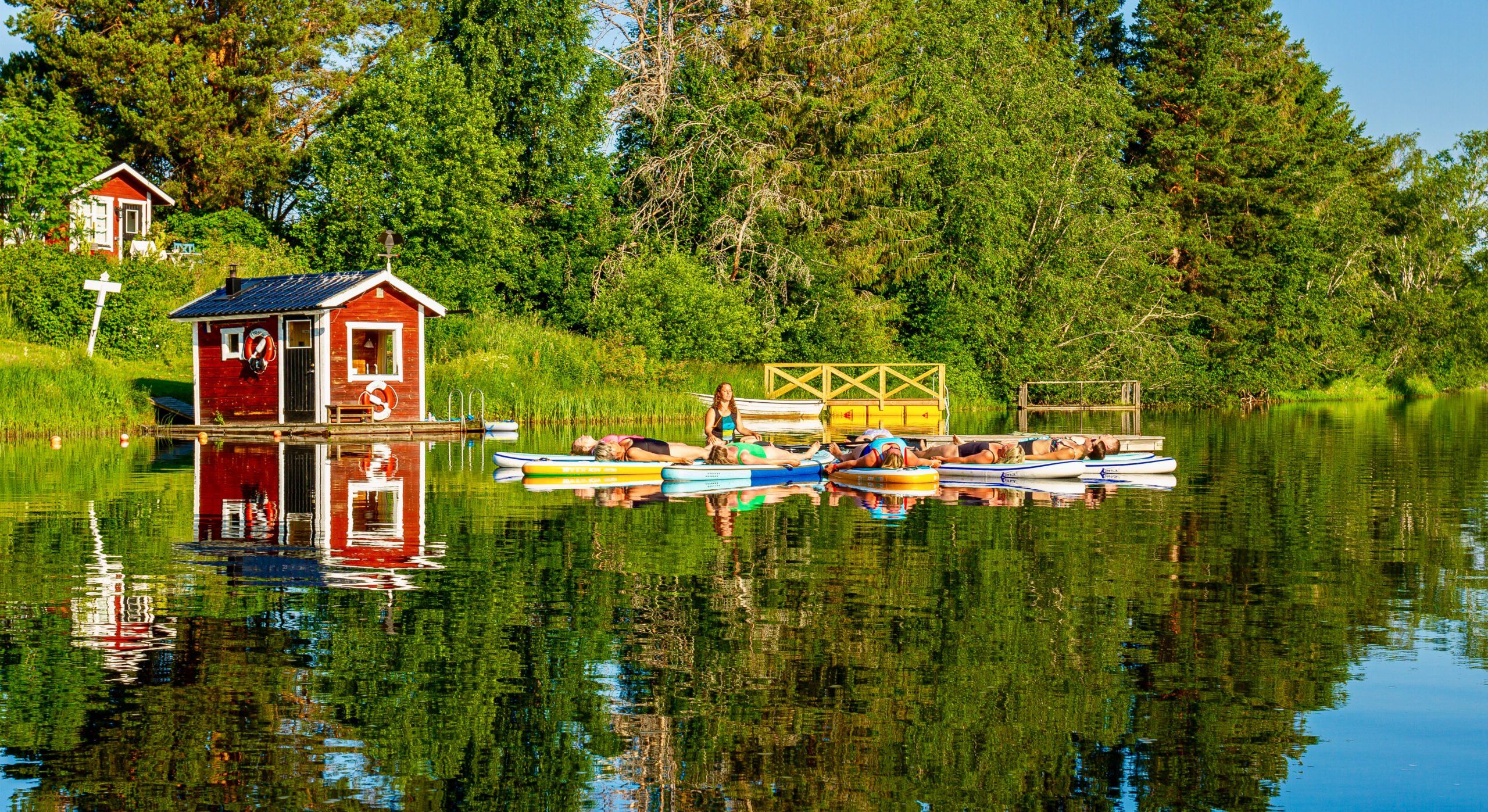 SUP-yoga på Flosjön: deltagare vilar på sina brädor intill bastun vid bryggan, med skogen speglad i vattnet.