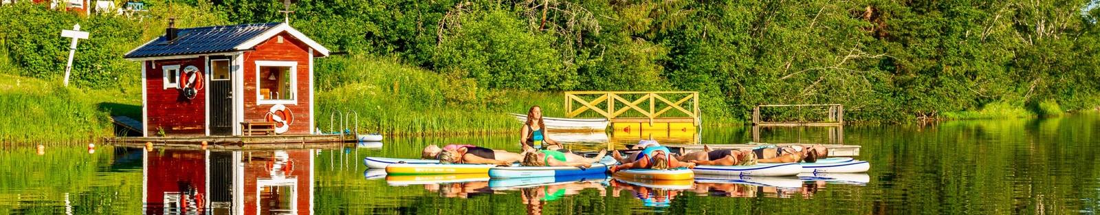 SUP-yoga på Flosjön: deltagare vilar på sina brädor intill bastun vid bryggan, med skogen speglad i vattnet.