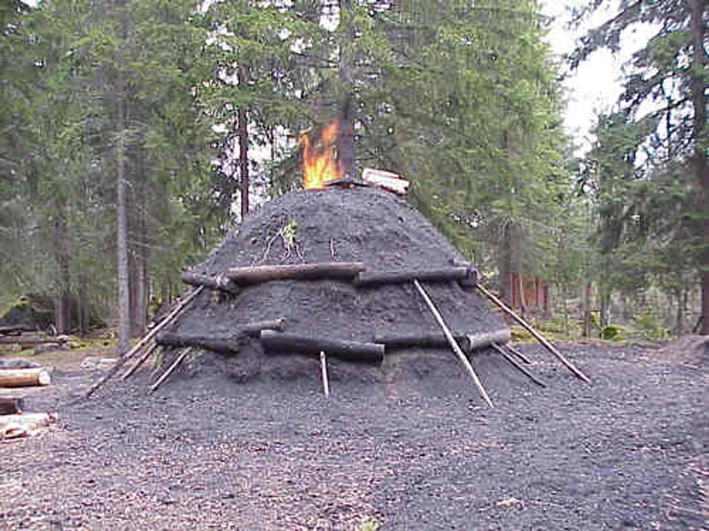 En kolmila i granskogen vid Kvarna: en låg kupol täckt av jord och kolstybb, stöttad av stänger, med lågor ur toppen.