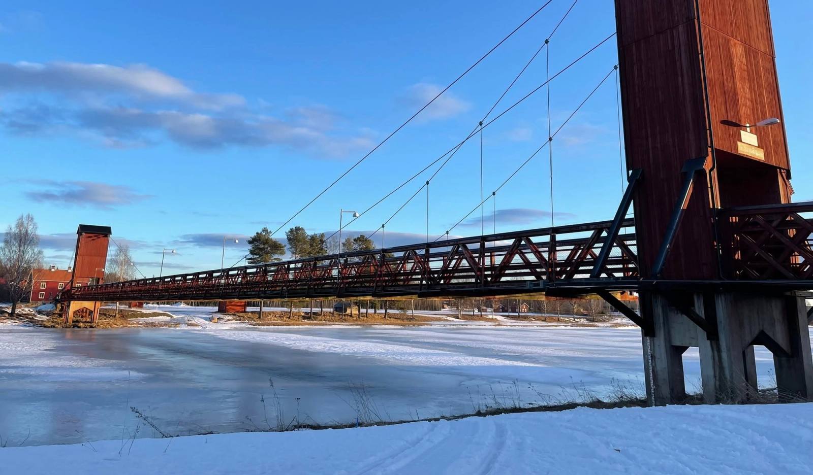 Hängbron över den isbelagda älven, med sitt röda brotorn mot blå himmel.