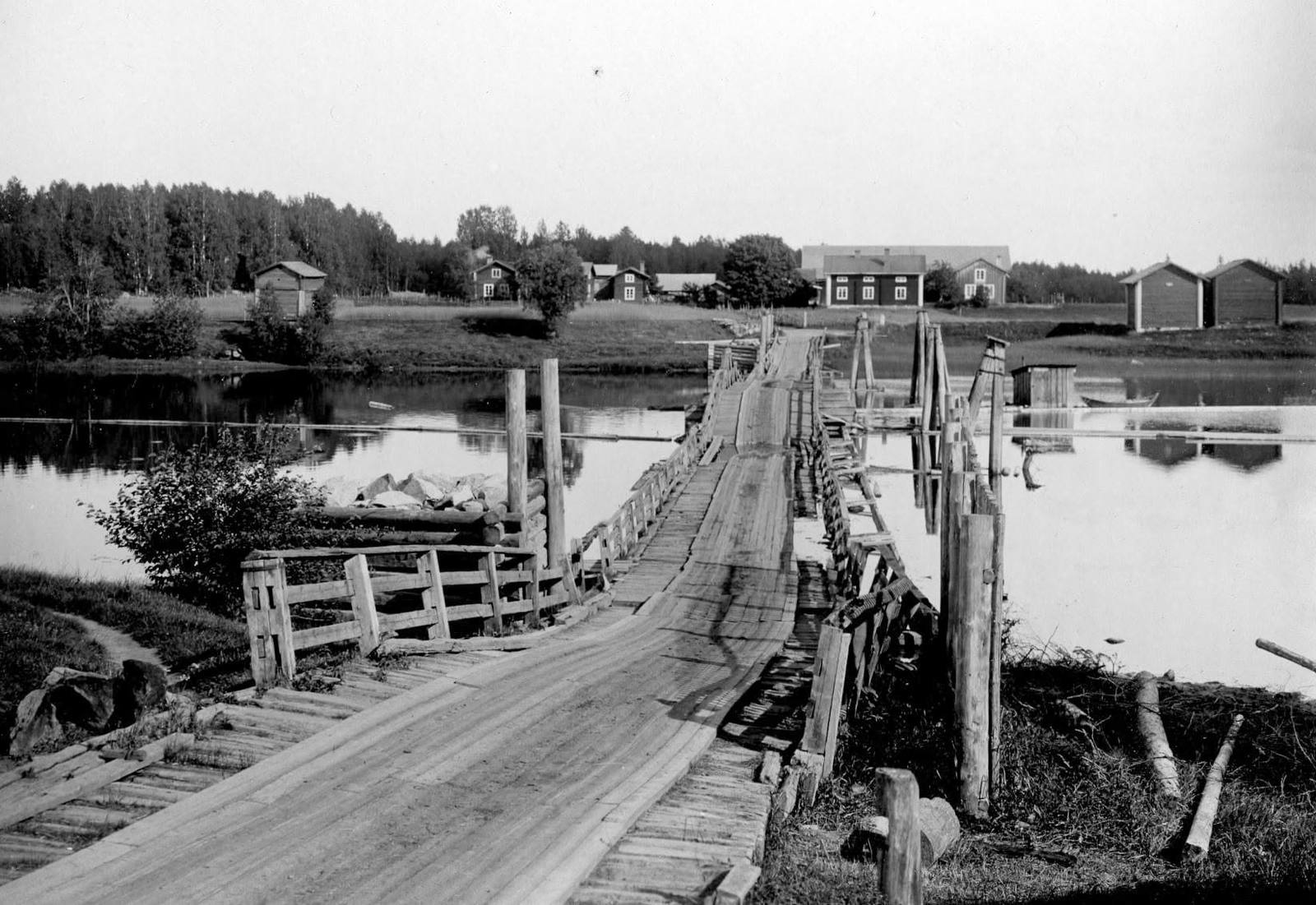 Gammalt svartvitt fotografi av en flottbro över älven, med byns hus och båthus på andra sidan.