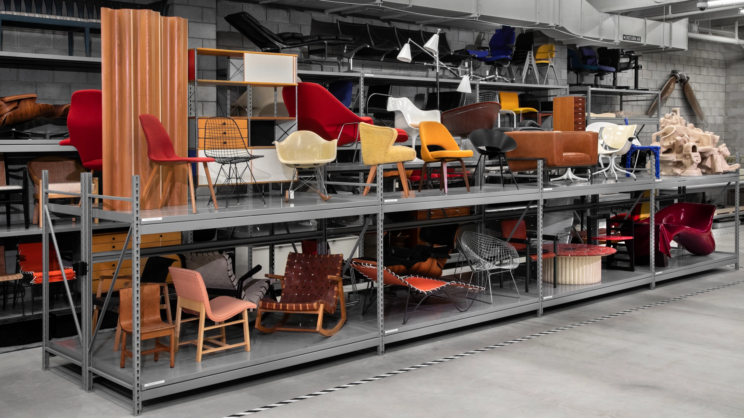 Metal shelves at the Cranbrook Art Museum filled with Eames designs such as the Folding Screen-Wood, a wire chair, and an RAR Rocking Chair, among others.