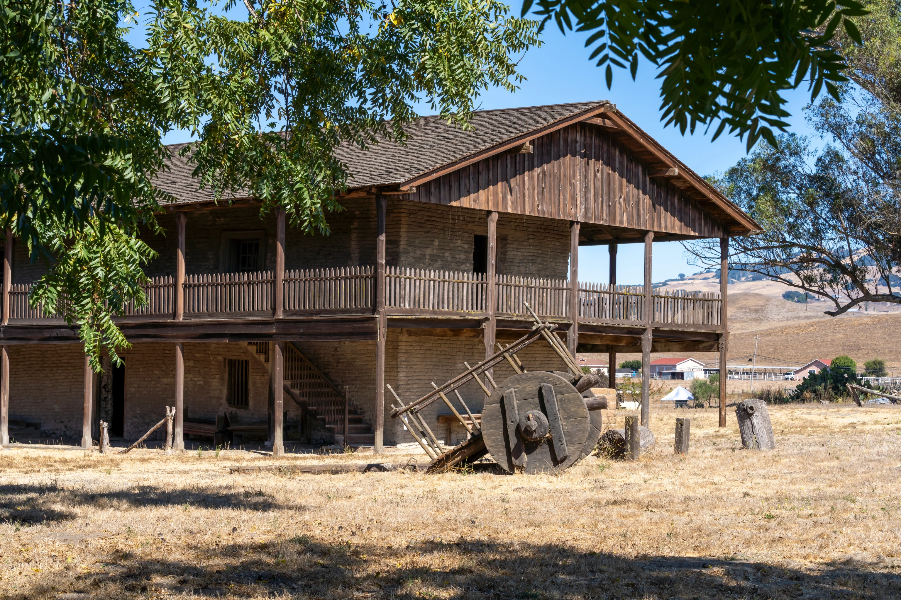 Exterior of Petaluma Adobe State Historic Park, showing the two-story adobe ranch house with wraparound wooden verandas and a historic millstone in the foreground.