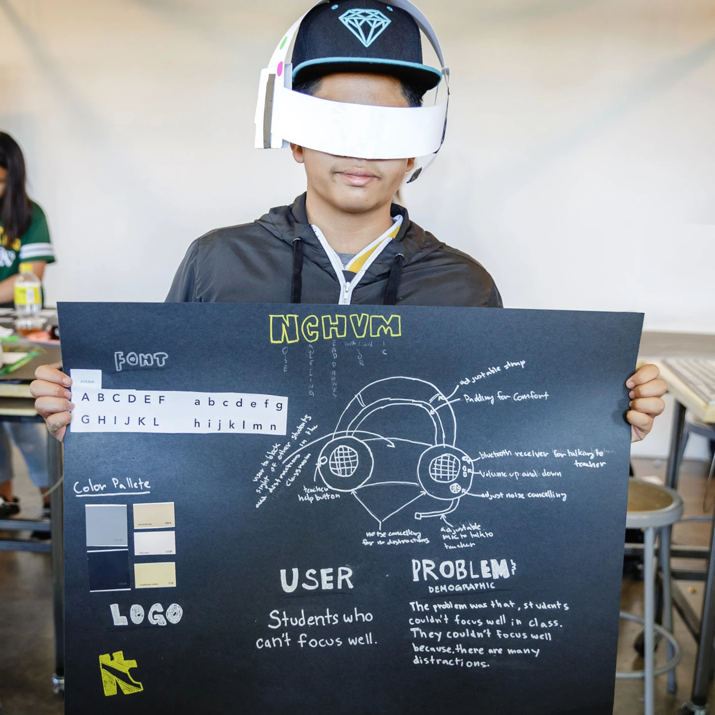 Inneract Project design student wearing a paper prototype and holding up a poster with details of his design solution.