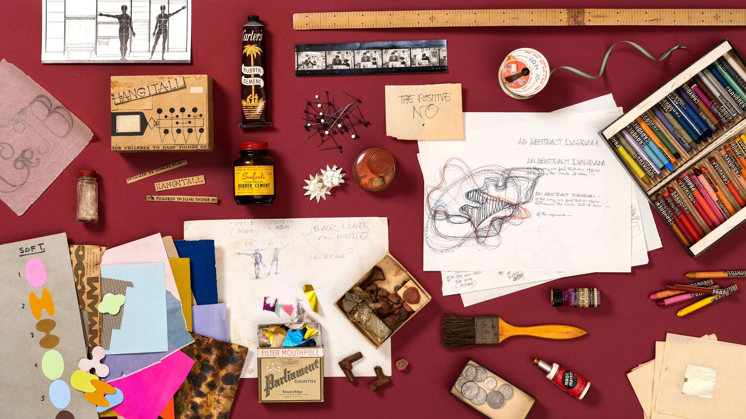 Various sketches and mock-ups created by artist and designer Ray Eames spread out on a table with some tools, and ephemera.