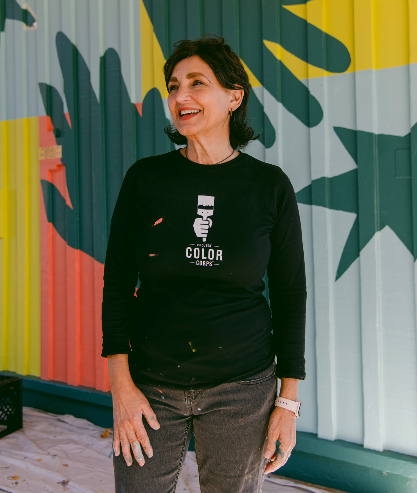 Laura Guido-Clark, standing to the side of a brightly colored mural painted by Project Color Corps.