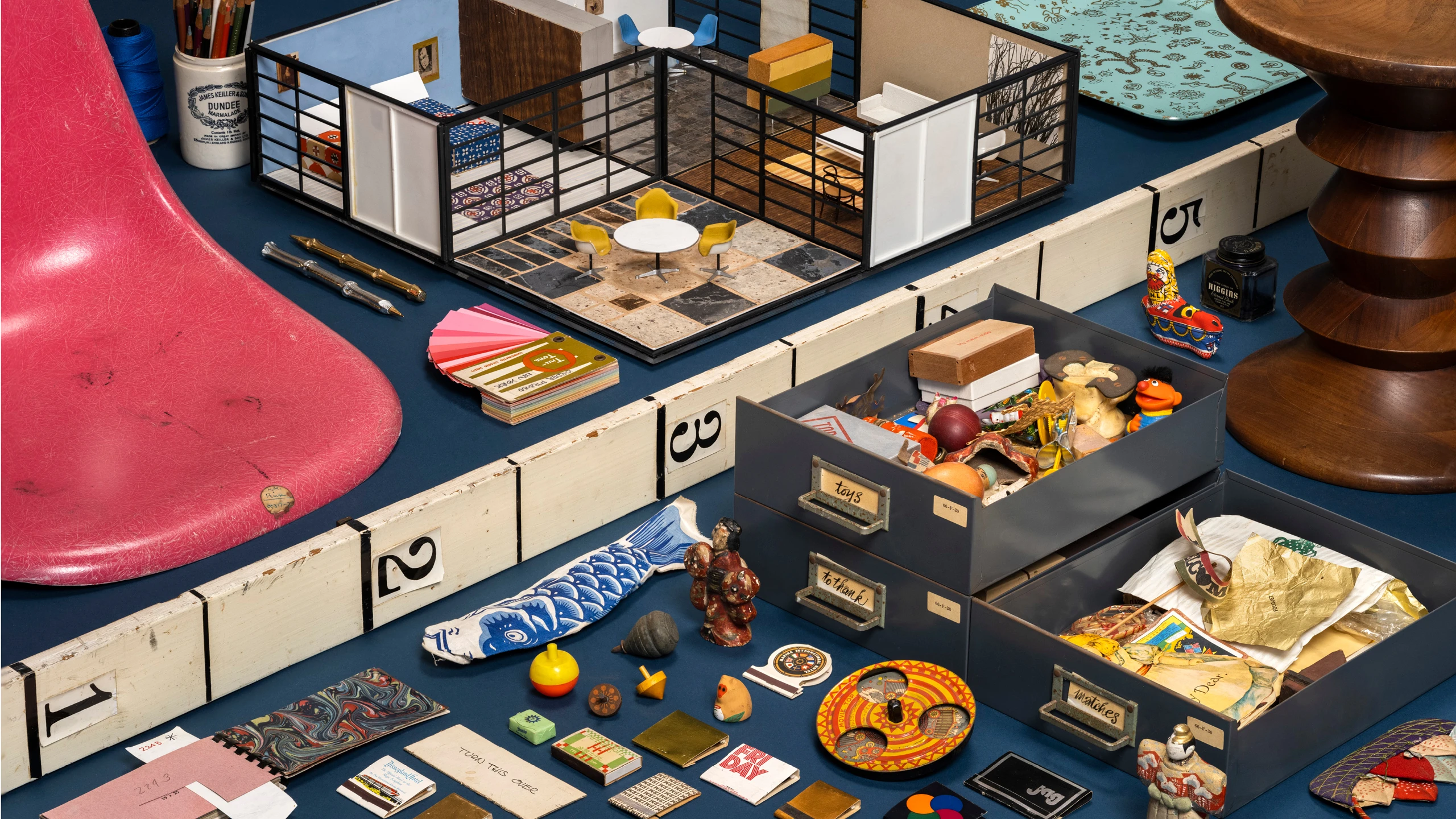 Eames designs such as the Revell Toy house model, walnut stool, shell chair, and various ephemera laid out on a table.
