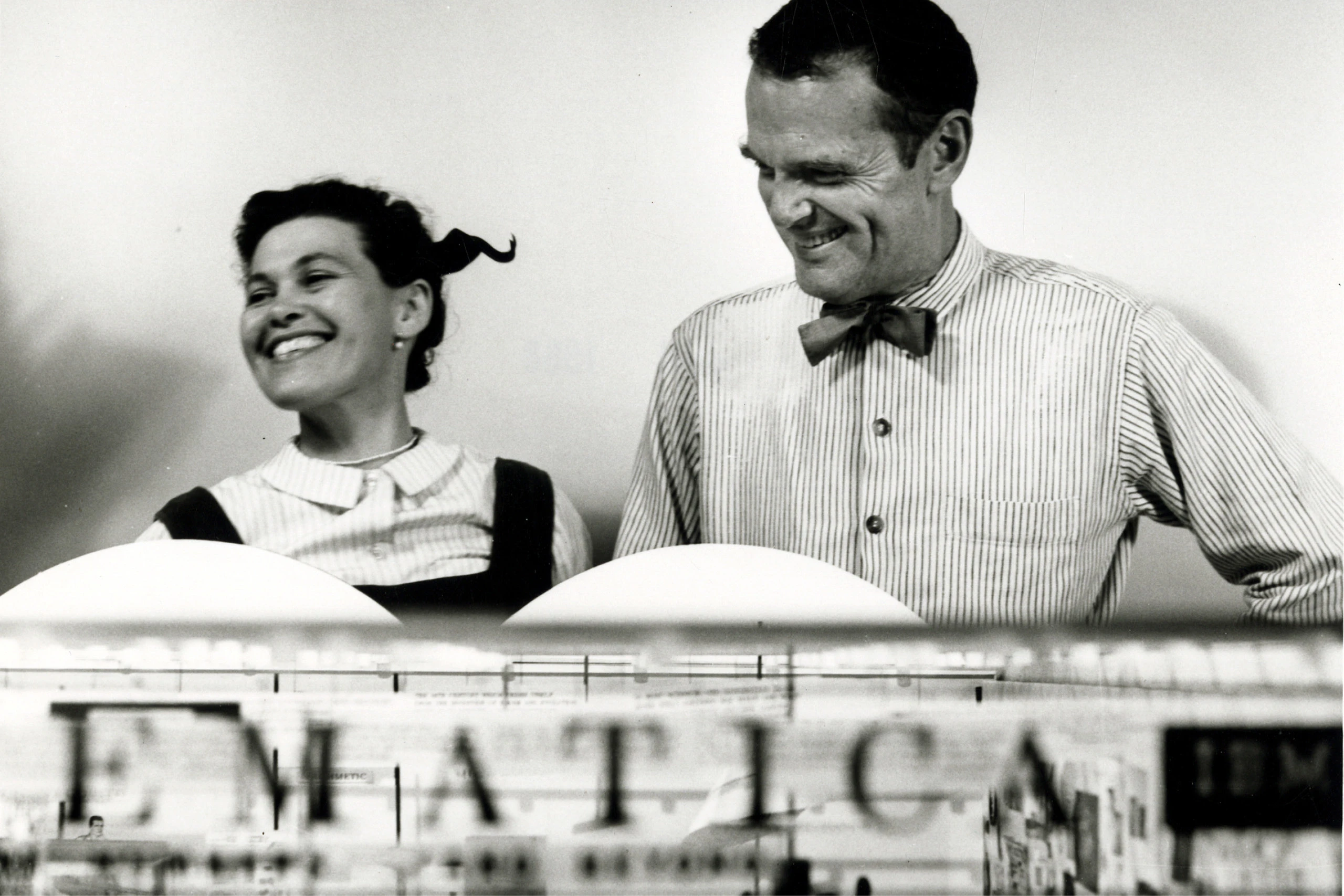 Ray and Charles Eames smiling while beside a scale model of the Mathematica Exhibition they designed.
