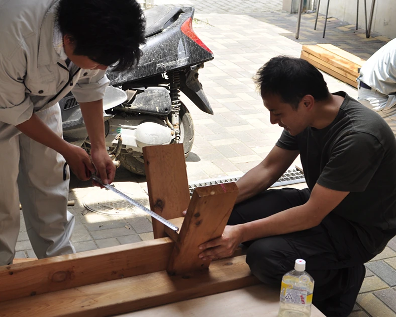 Ishinomaki Lab founder Keiji Ashizawa sitting down helping another man assemble a handmade wooden bench, Kazam! Magazine.