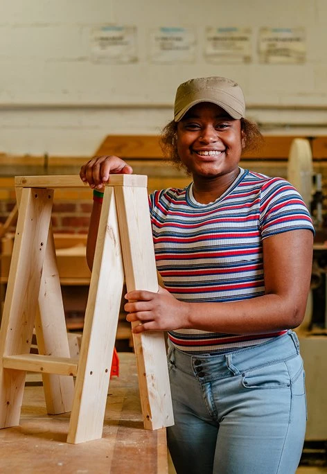 At Brightmoor Maker Space, a young woman stands in the workshop holding a handmade Ishinomaki wooden stool, Kazam! Magazine.
