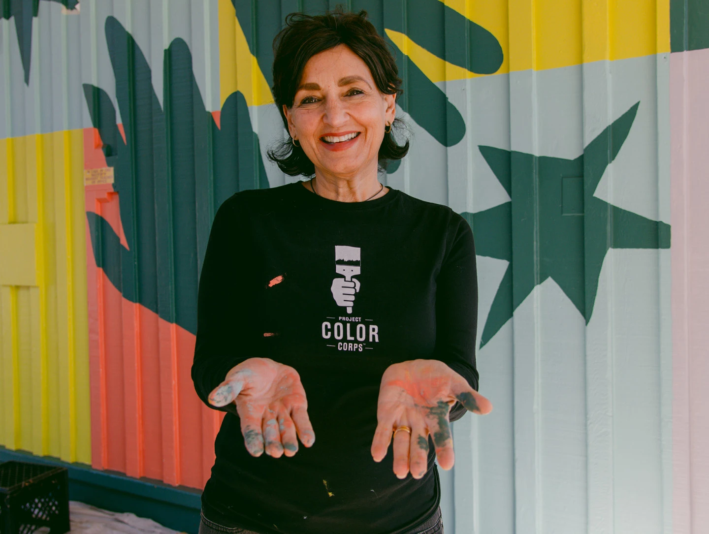 Laura Guido-Clark of Project Color Corps standing in front of a brightly colored mural with paint on her outstretched hands.
