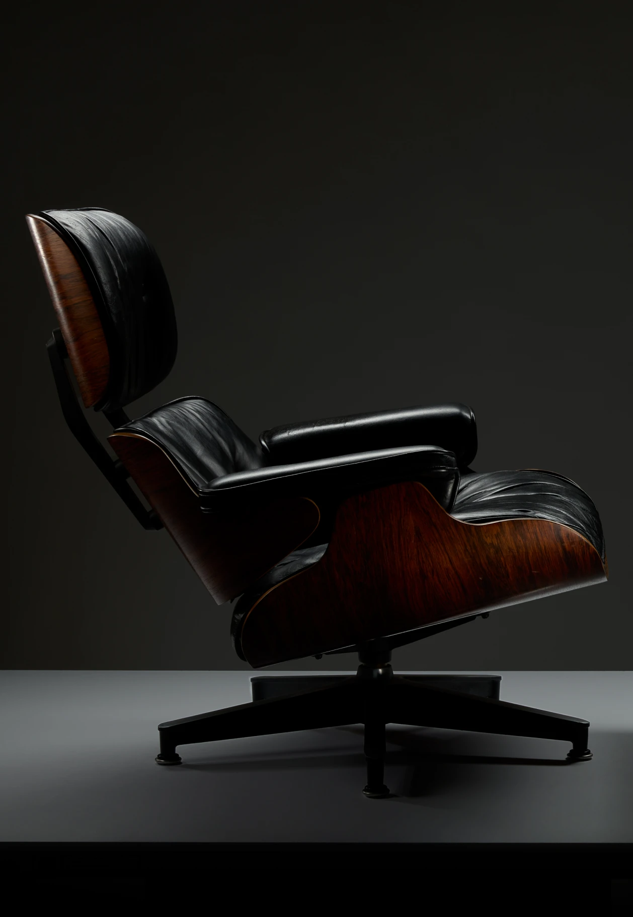 Side view of the Eames lounge chair highlighting the molded rosewood and black leather in dramatic lighting.