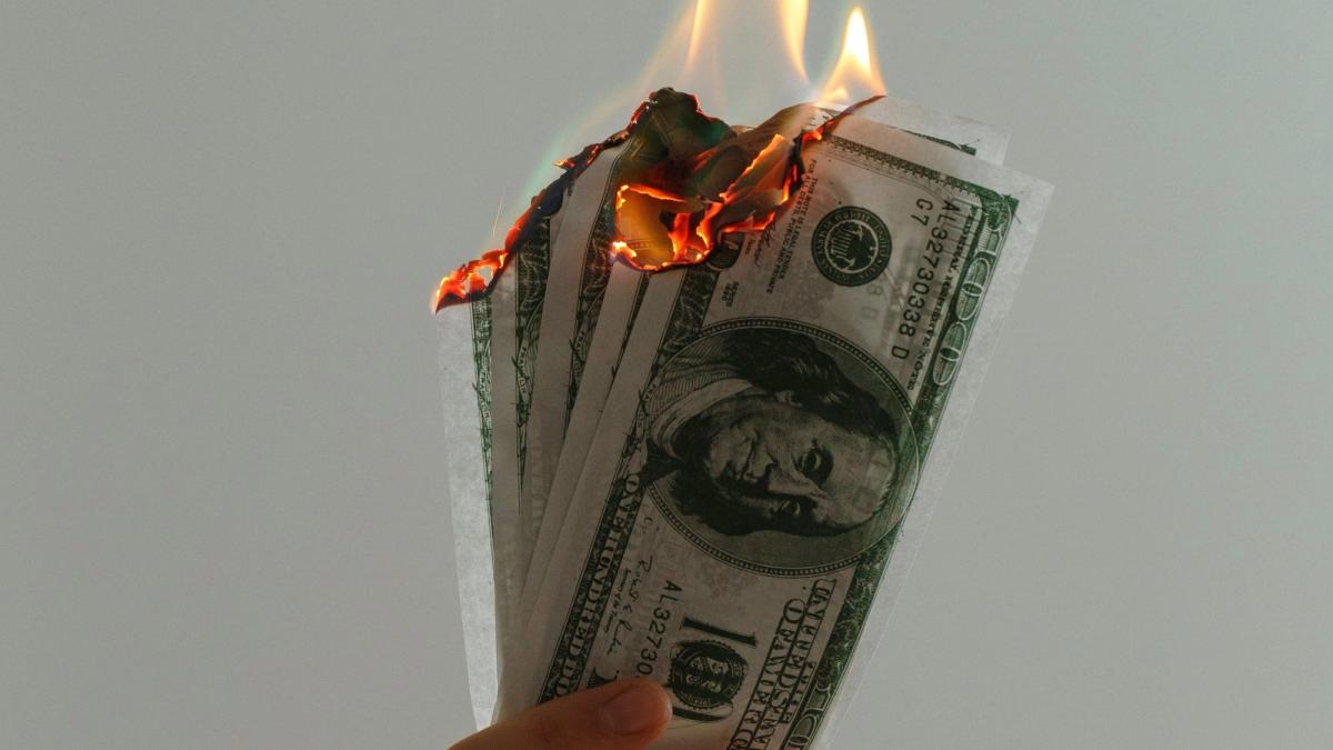A hand holds several burning hundred-dollar bills.