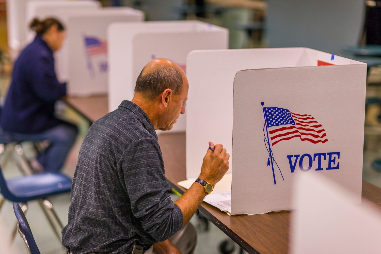 A voter sits at a desk with a ballot paper shielded by a partition bearing the U.S. flag and the word 'Vote.'