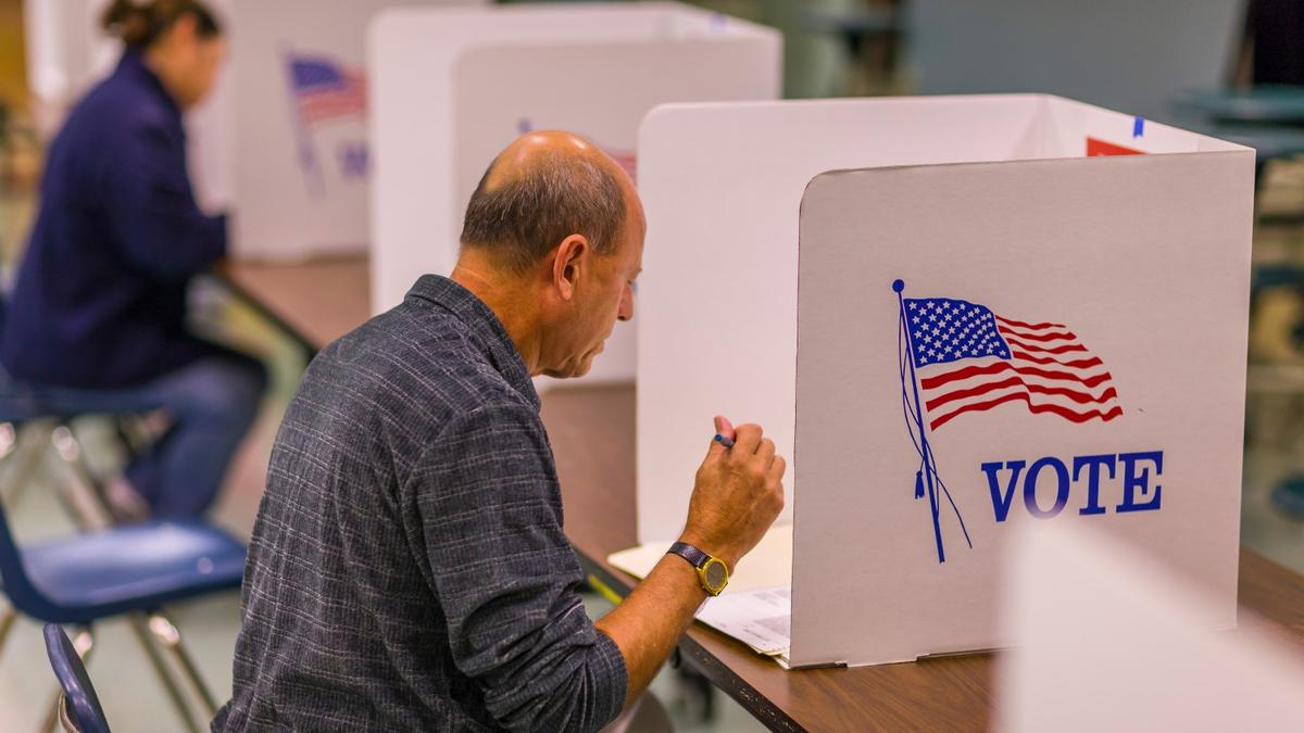 A voter sits at a desk with a ballot paper shielded by a partition bearing the U.S. flag and the word 'Vote.'