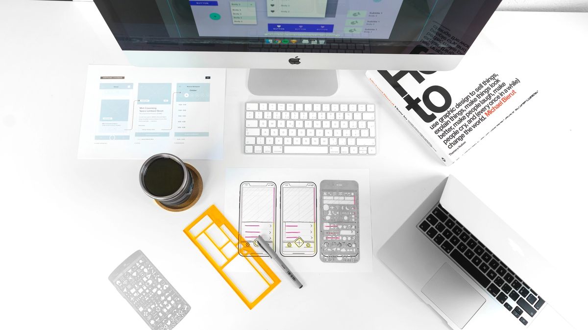 Bird's eye view of a desk with a Mac computer, keyboard, MacBook, papers, stencils, a design book and a cup of coffee.