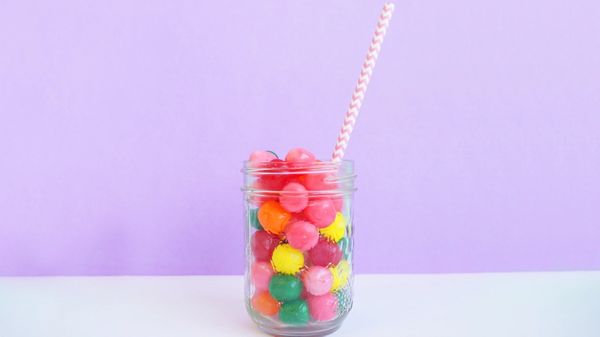 Jar of coloured sweets with a striped straw in, against a white and purple background.