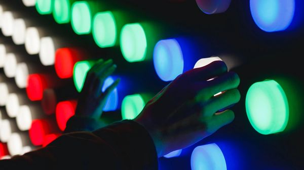 Hands pressing large colourful light-up buttons, on a black board.