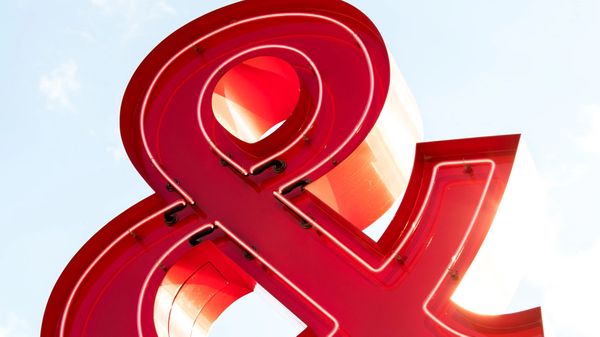 Large, red, metal ampersand sign on a pole, against a backdrop of a blue sky with scattered clouds.