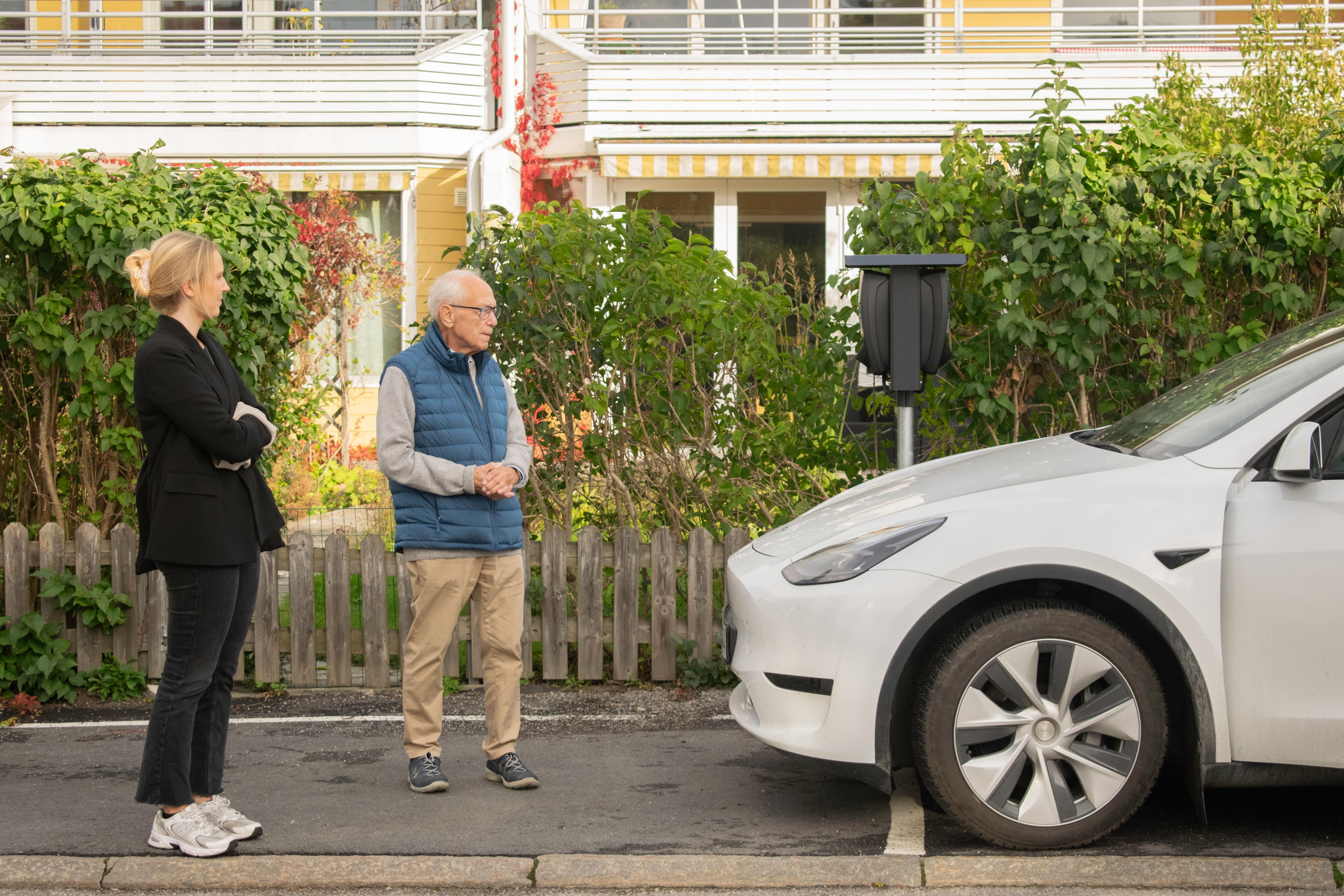 En mann og en dame som står utenfor et borettslag ved siden av en elbil og en ladeboks for elbillading