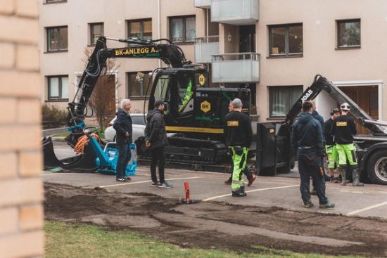 Bilde av en parkeringsplass med gravmaskin og mennesker som arbeider utenfor et leilighetskompleks