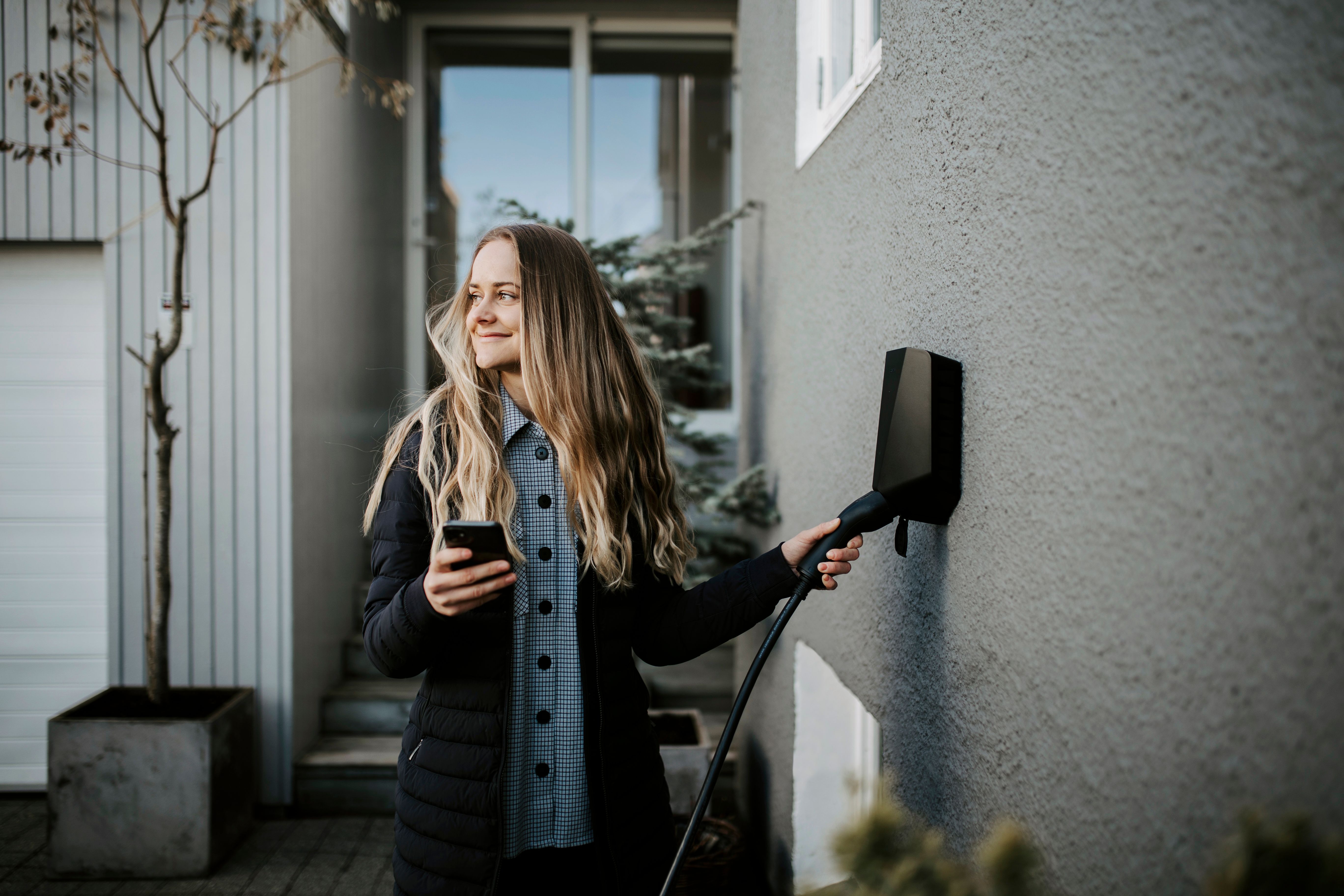 Smilende kvinne med langt hår og mobil i hånden strekker seg etter svart elbillader på veggen.