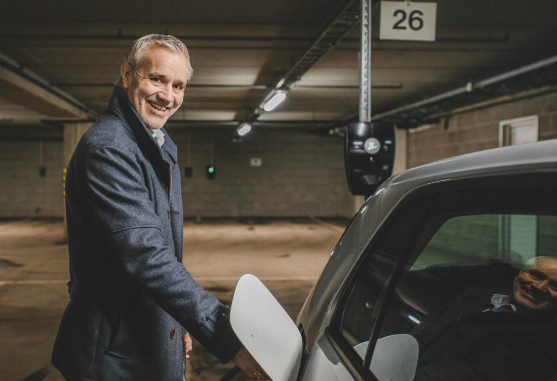 Foto av mann som står ved siden av bilen sitt i ett garasjeanlegg og smiler til kameraet