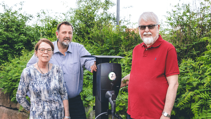 Bilde av en dame og 2 menn utendørs ved siden av en ladestolpe for elbillading